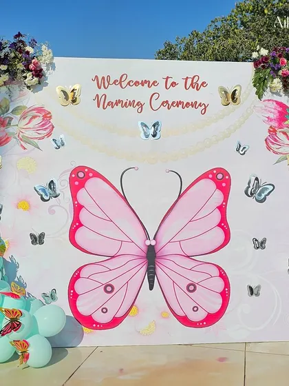 A fun and interactive photo booth for the naming ceremony. The backdrop features a large pink butterfly, a balloon arch, and smaller silver butterflies for a perfect picture spot.