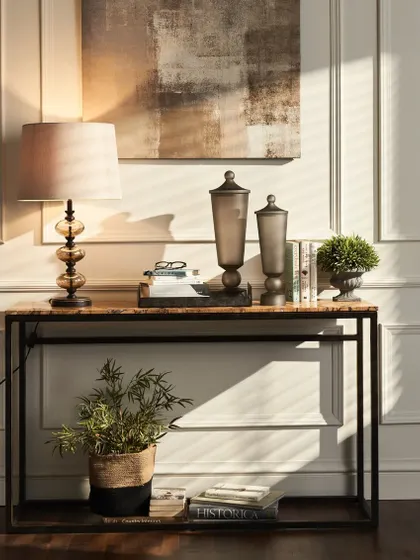 A console table styled with lamps, books, and decorative jars. The dramatic shadows from the window blinds add depth and mood to this elegant interior vignette.
