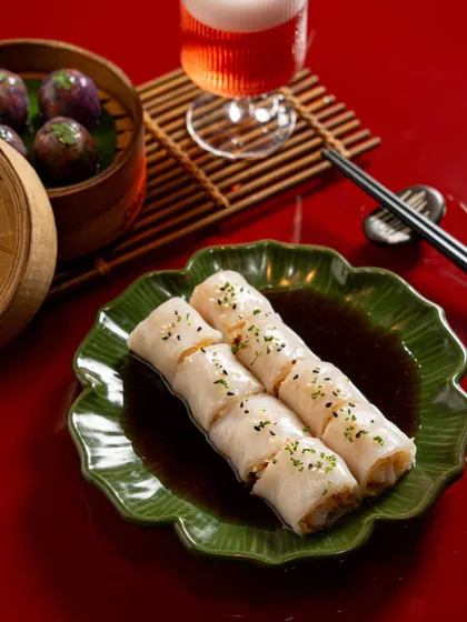 A shot of Cheung Fun (rice noodle rolls) in a savory sauce, paired with a cocktail. The composition uses a strong red background and focused lighting to make the dish pop.