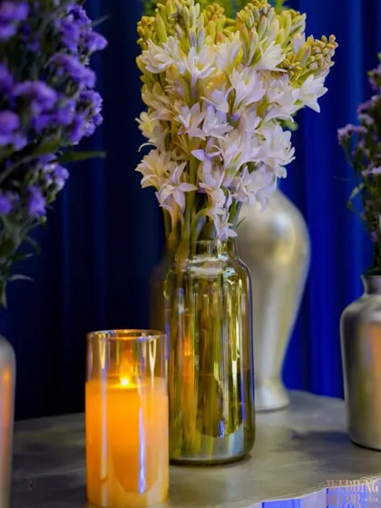 The soft glow of a candle next to a vase of fragrant tuberoses, set against the rich blue velvet backdrop. These small details add warmth and sensory richness to the space.
