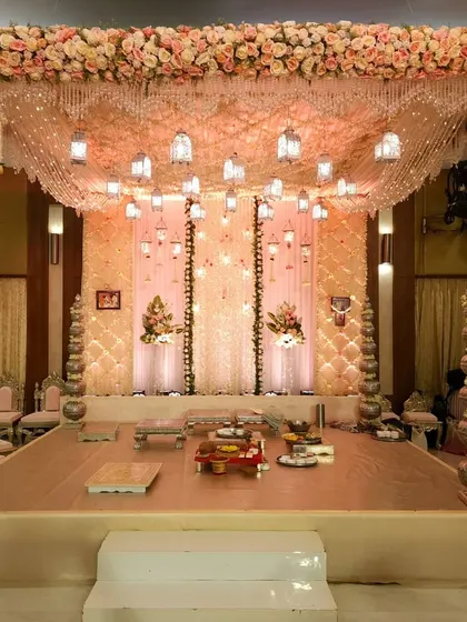 This is a beautiful pastel wedding mandap, featuring a canopy of draped fabric and hanging lanterns. The structure is supported by ornate silver pillars, and the entire stage is decorated with a thick border of pink and white roses for a classic, romantic feel.