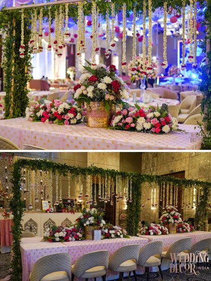 A long view of a buffet or bar setup, decorated with a canopy of greenery and hanging floral strings. The tables are adorned with beautiful arrangements of red, pink, and white flowers.