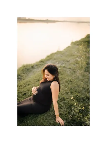 A wider perspective of the serene lakeside scene, showing the expecting mother enjoying a peaceful moment in nature. The calm water and soft light create a tranquil atmosphere.