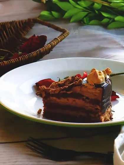 A slice of rich chocolate pastry on a white plate, styled with rose petals. The natural, soft light from the side creates a gentle and romantic mood.