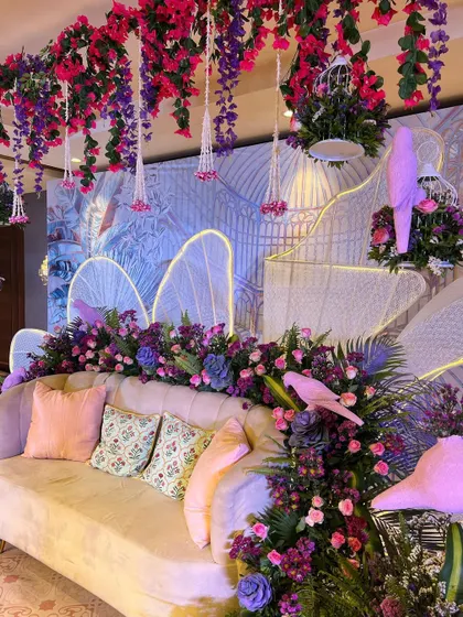 The main seating area of the 'Illuminated Garden', with a plush sofa surrounded by glowing wings, hanging florals, and artistic props.