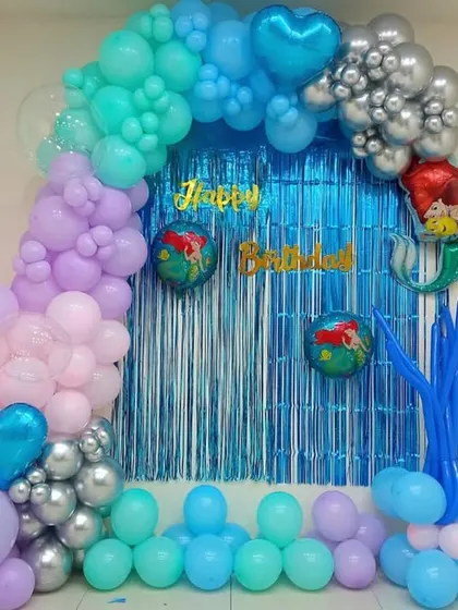 A simple mermaid-themed decoration for a home party. This setup uses a blue foil curtain for a backdrop and a colorful balloon arch with mermaid foil balloons.