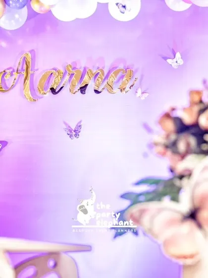 The birthday girl's name in gold lettering on a purple-lit wall with fluttering butterflies creates a simple yet elegant focal point.