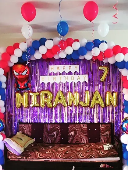 A classic Spiderman birthday party setup, perfect for a superhero fan's seventh birthday. The decoration features a red, white, and blue balloon arch over a couch, set against a purple shimmer curtain. A Spiderman foil balloon and the name "NIRANJAN" in gold letters make it clear who the hero of the day is.