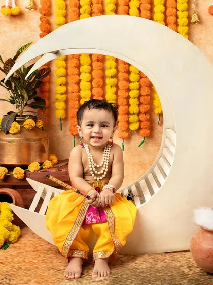 A smiling baby dressed as Krishna sits happily on the moon prop, ready for his portrait.