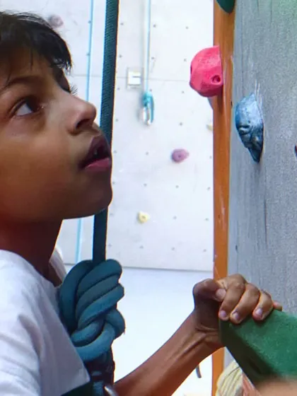 A young climber looks up, planning his route. Our programs encourage kids to think strategically and build mental focus alongside physical strength.