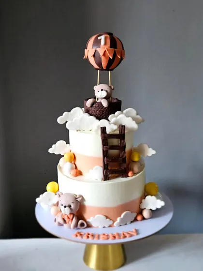 A lovely hot air balloon themed cake for a first birthday. This two-tiered design features teddy bears, edible clouds, and a hot air balloon topper, all in a soft, neutral color palette.