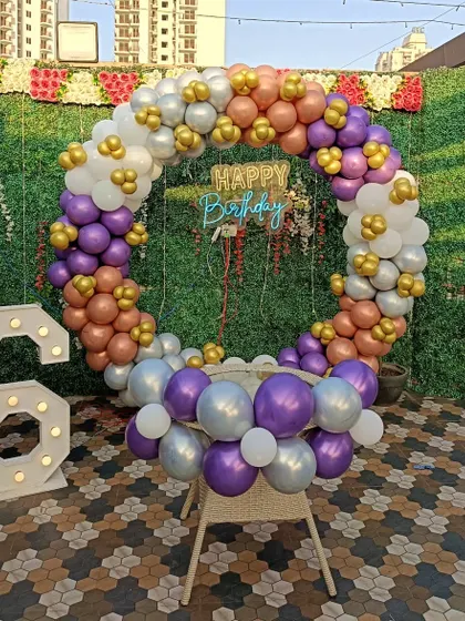 An outdoor birthday setup with a multi-color balloon ring against a green wall. The decoration includes a 'Happy Birthday' neon sign and a large light-up number '6'.