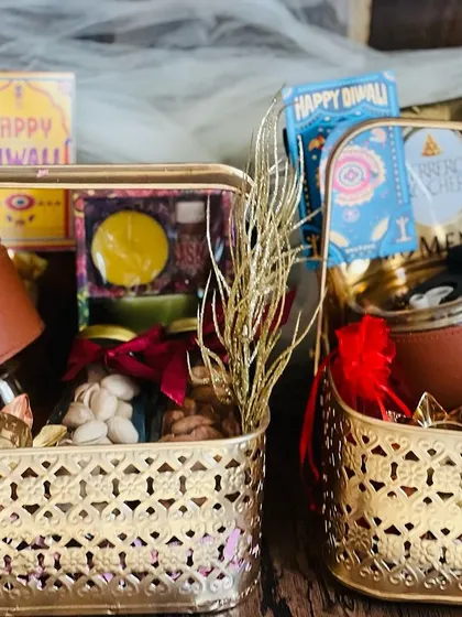 Two golden Diwali baskets side-by-side, filled with jars of nuts, festive cards, and decorative items.