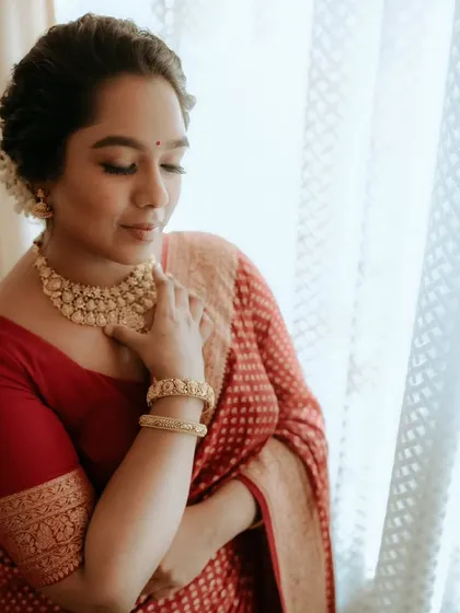 A bride in a red saree looks down, a quiet moment captured by the window, highlighting her jewelry and serene expression.