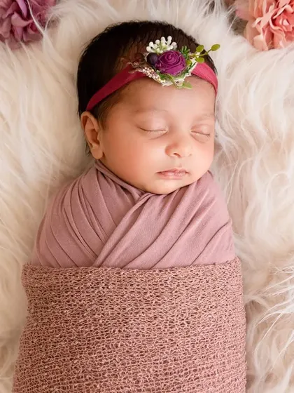 A close-up of a sleeping newborn swaddled on a white fur rug, wearing a delicate floral headband.