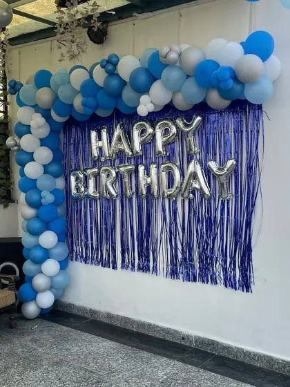 An outdoor birthday backdrop with a blue shimmer curtain and a blue, grey, and white balloon arch.