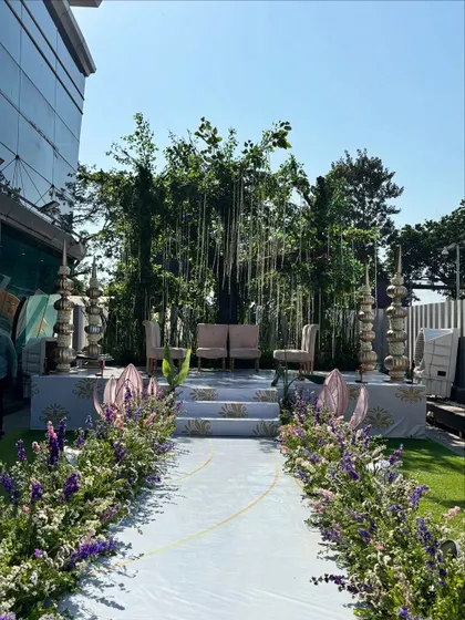 A beautiful outdoor aisle for a Varmala ceremony. The path is lined with purple and white wildflowers, leading to a stage set against a natural backdrop of lush green trees, creating a serene and romantic setting.