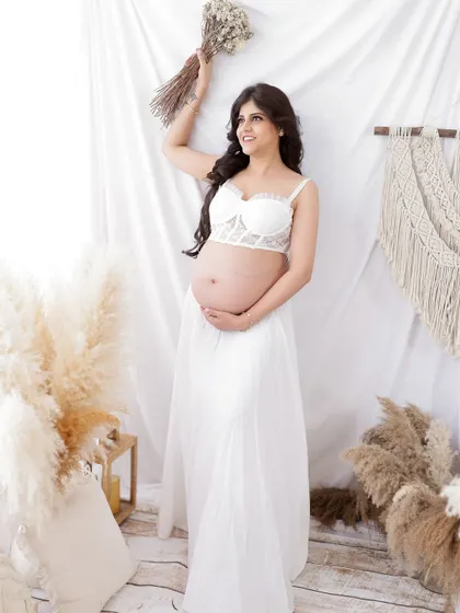 A joyful and free-spirited portrait. The mom-to-be is dressed in a white boho-style outfit, holding a bouquet of dried flowers with a happy expression.