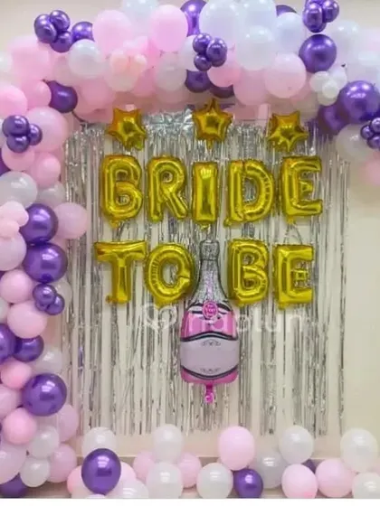 This backdrop uses a silver shimmer curtain and a large balloon arch in purple, pink, and white. The 'BRIDE TO BE' is spelled out in large gold foil balloons, creating a fun and celebratory look.