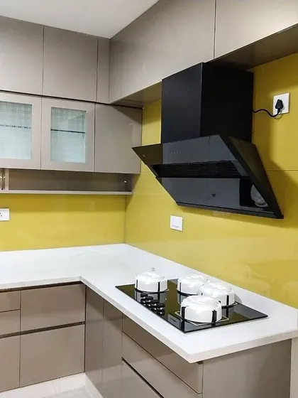 A contemporary L-shaped kitchen featuring neutral beige cabinets and a bright yellow lacquered glass backsplash that adds a pop of color. The design includes a black chimney and frosted glass upper cabinets for a clean and modern look.