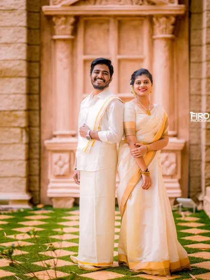 An elegant portrait of a couple in classic white and gold Kerala-style attire. Their timeless outfits look stunning against the grand architectural backdrop.