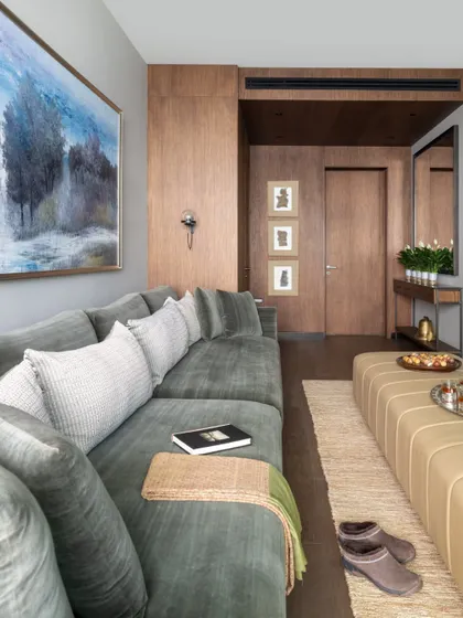 The family lounge in the DLF Camellias apartment is designed for ultimate comfort. A deep, plush sectional sofa in a calming green-grey chenille is perfect for family movie nights, complemented by a large leather ottoman and a serene landscape painting.