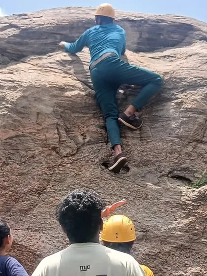 An instructor guides a student's foot placement while others spot from below during a bouldering exercise.