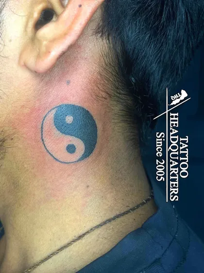 A solid black Yin and Yang symbol tattooed behind the ear, representing balance, harmony, and the duality of the universe.