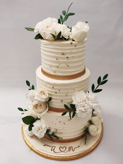 We design the picture of wedding day elegance with this royal white floral cake. Two tiers of textured buttercream are wrapped in gold ribbon and adorned with bouquets of fresh white roses and carnations, creating a timeless and sophisticated wedding centerpiece.