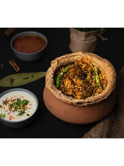 A clay pot of handi biryani, styled with raita and salan against a dark, textured background. The use of traditional serving ware and spices like cinnamon and bay leaf enhances the authentic appeal.