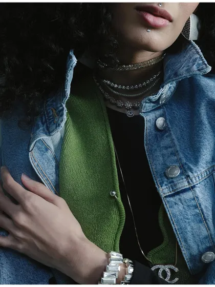 A close-up detail shot of layered denim, knitwear, and delicate necklaces. This image highlights the texture and detail of the styling in a moody, intimate composition.