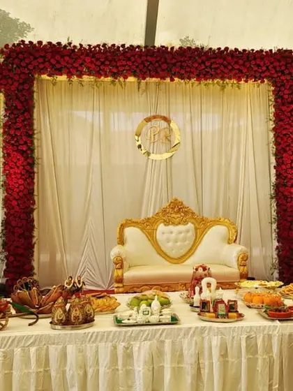 A classic engagement stage framed by three arches of dense red roses. The setup includes a traditional white and gold sofa and a table laden with ceremonial offerings.