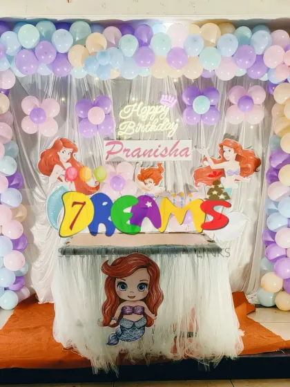 A simple and sweet mermaid theme for a home party. This setup uses a pastel balloon arch over a white curtain backdrop with Ariel character cutouts and a themed table skirt.