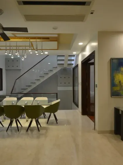 The first-floor dining room at Gupta Residence is a modern, elegant space. A glass-top table with sleek olive-green chairs is highlighted by pendant lighting, with a floating staircase forming a striking backdrop.