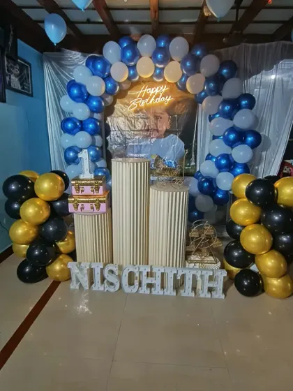 We travel for outside decorations, like this first birthday event in Hoovinamadu. The setup includes a blue and white balloon arch, gold and black balloon columns, and the name 'NISCHITH' in letters.