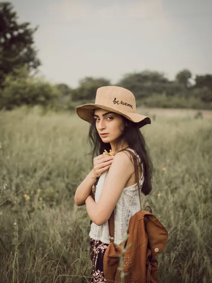 A vintage-inspired portrait in a green field. The soft, natural lighting and the styling with the hat and backpack create a story of a traveler, giving the image a narrative quality.