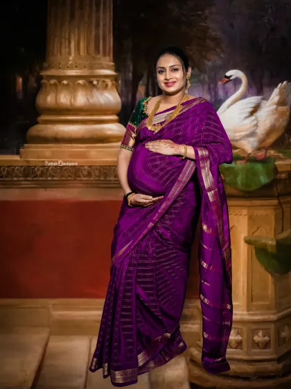 A regal and artistic portrait. The expecting mother in her purple saree looks like a painting, standing beside a classic pillar and a graceful swan.