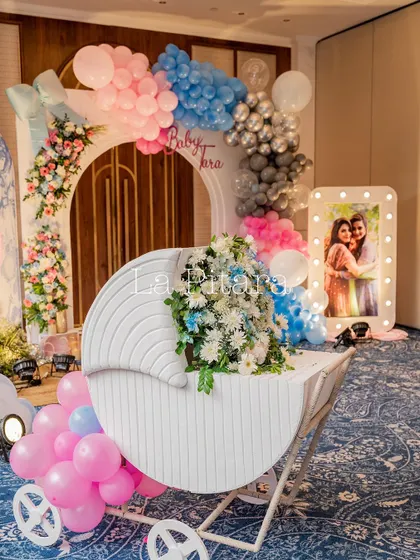 A modern baby cradle filled with fresh flowers, set against a balloon arch and an illuminated photo of the happy couple.