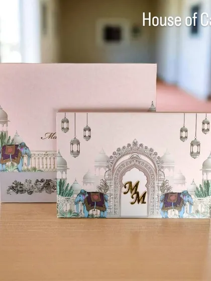 This pastel pink invitation features a palace-inspired design with elephants and hanging lanterns, complete with a custom gold monogram for a personal touch.