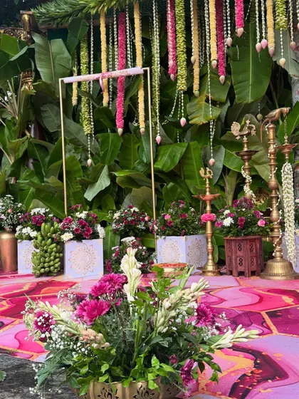 A close-up of the traditional decor elements, including hanging floral strings and brass planters, against a backdrop of banana leaves.