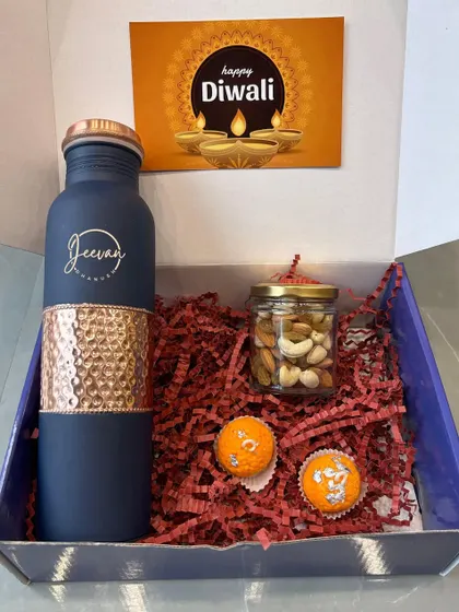 A personalized Diwali gift box featuring a blue and hammered copper bottle engraved with the name "Jeevan". The hamper also includes a jar of dry fruits and mithai-shaped candles.