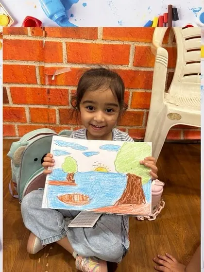 A young girl proudly holds up her beautiful landscape drawing. We teach various techniques, from coloring with crayons to painting.