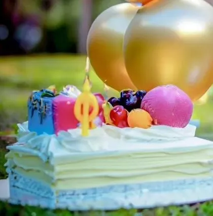 A colorful birthday cake topped with fruit, a candle, and golden balloons, ready for an outdoor celebration.