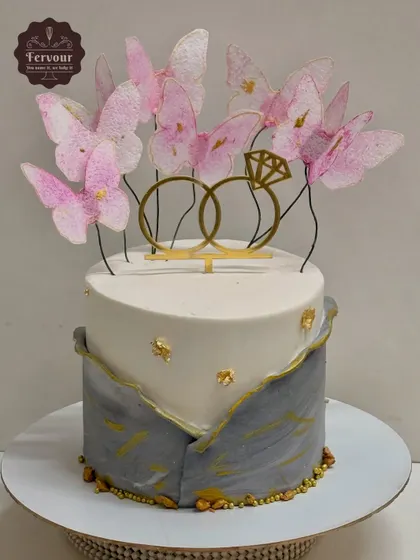 An elegant two-tier engagement cake featuring a grey marble effect on the bottom tier and delicate pink wafer paper butterflies. The gold ring topper and gold leaf accents add a touch of sophistication.