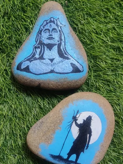 A set of two river stones painted with different depictions of Lord Shiva. One shows the meditative Adiyogi, and the other shows his powerful silhouette against the moon.
