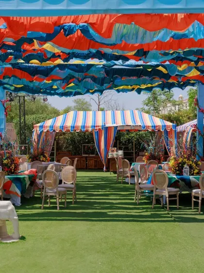 A wide view of a carnival-themed wedding event. The colorful striped tents and wavy fabric ceiling in shades of blue, orange, and yellow create a festive and energetic atmosphere.