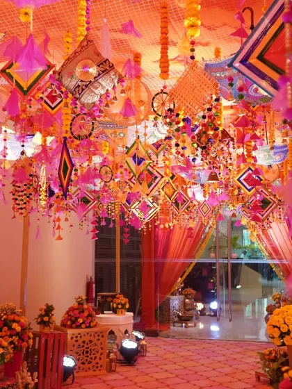 A stunning ceiling treatment for a Mehendi ceremony. The entire ceiling is covered with colorful hanging phulkari and dreamcatcher-style ornaments, creating an immersive and festive canopy of color.