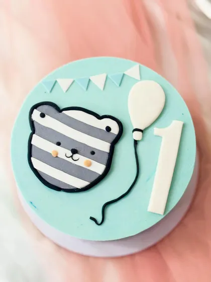 A minimalist first birthday cake with a cute striped teddy bear face. The simple design on a pale blue cake is modern and sweet.