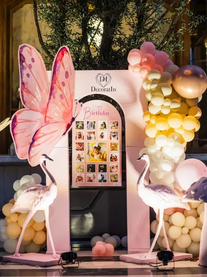 A memory lane display for a first birthday, featuring a grid of the baby's monthly photos. It's flanked by whimsical flamingo props and a giant butterfly, making it a heartwarming and beautiful corner.