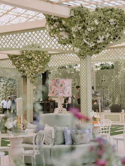 A lounge area designed under a custom pergola with heart-shaped floral installations on the pillars. The mix of ghost chairs and plush sofas with a custom-printed lampshade creates a whimsical and stylish corner.